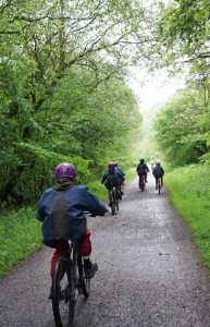 Photo taken from behind shows school children riding bikes along a green bushy stretch of the Monsal Trail